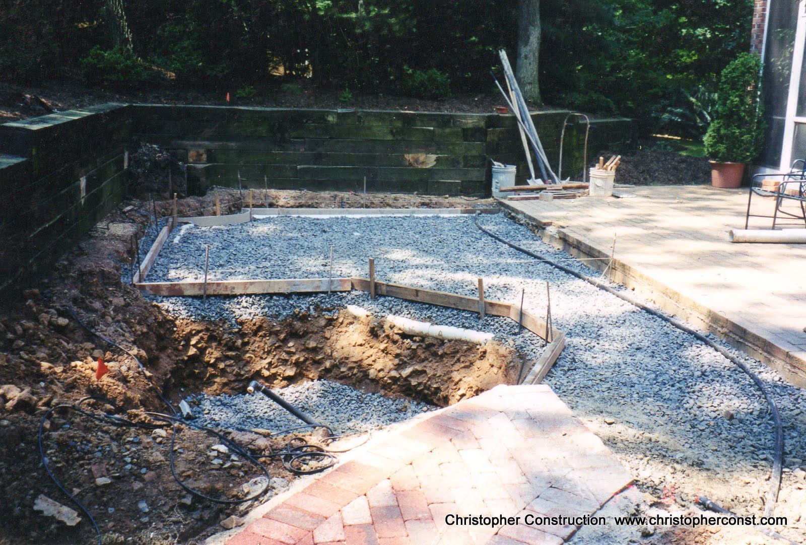 Empty backyard pool under construction with gravel, concrete edges, and a wooden deck nearby
