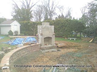 Outdoor stone fireplace under construction in a backyard, with stacked brick wall and tools nearby