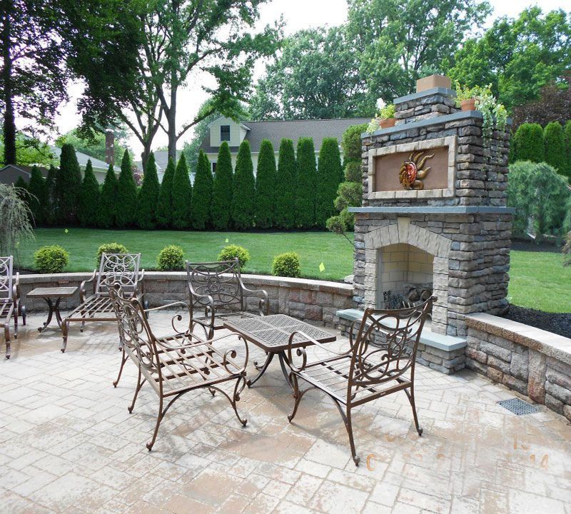 Stone patio with wrought-iron furniture and a brick outdoor fireplace in a landscaped backyard