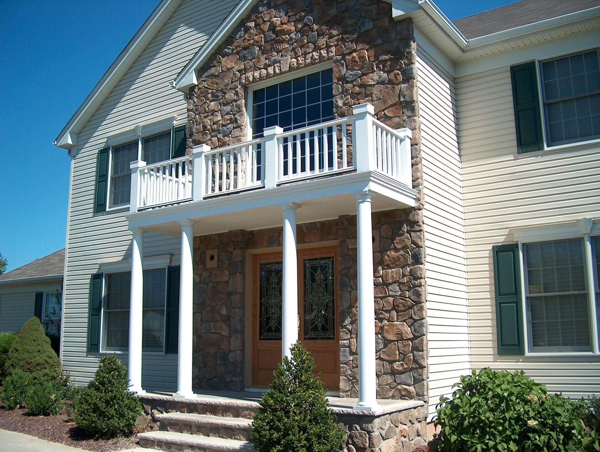 Two-story house with stone facade, white balcony, and front porch under a bright blue sky
