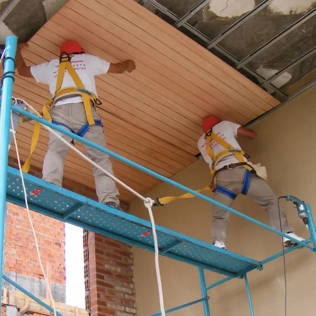Dos trabajadores con cascos rojos y arneses de seguridad pintan un techo sobre un andamio azul