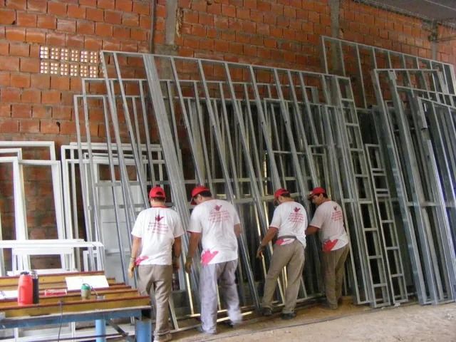 Trabajadores transportando marcos metálicos de ventanas dentro de un taller con paredes de ladrillo