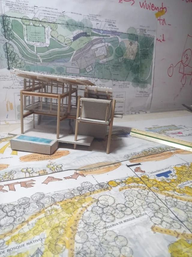 Maqueta arquitectónica sobre un escritorio, con planos del terreno y un mapa en la pared al fondo