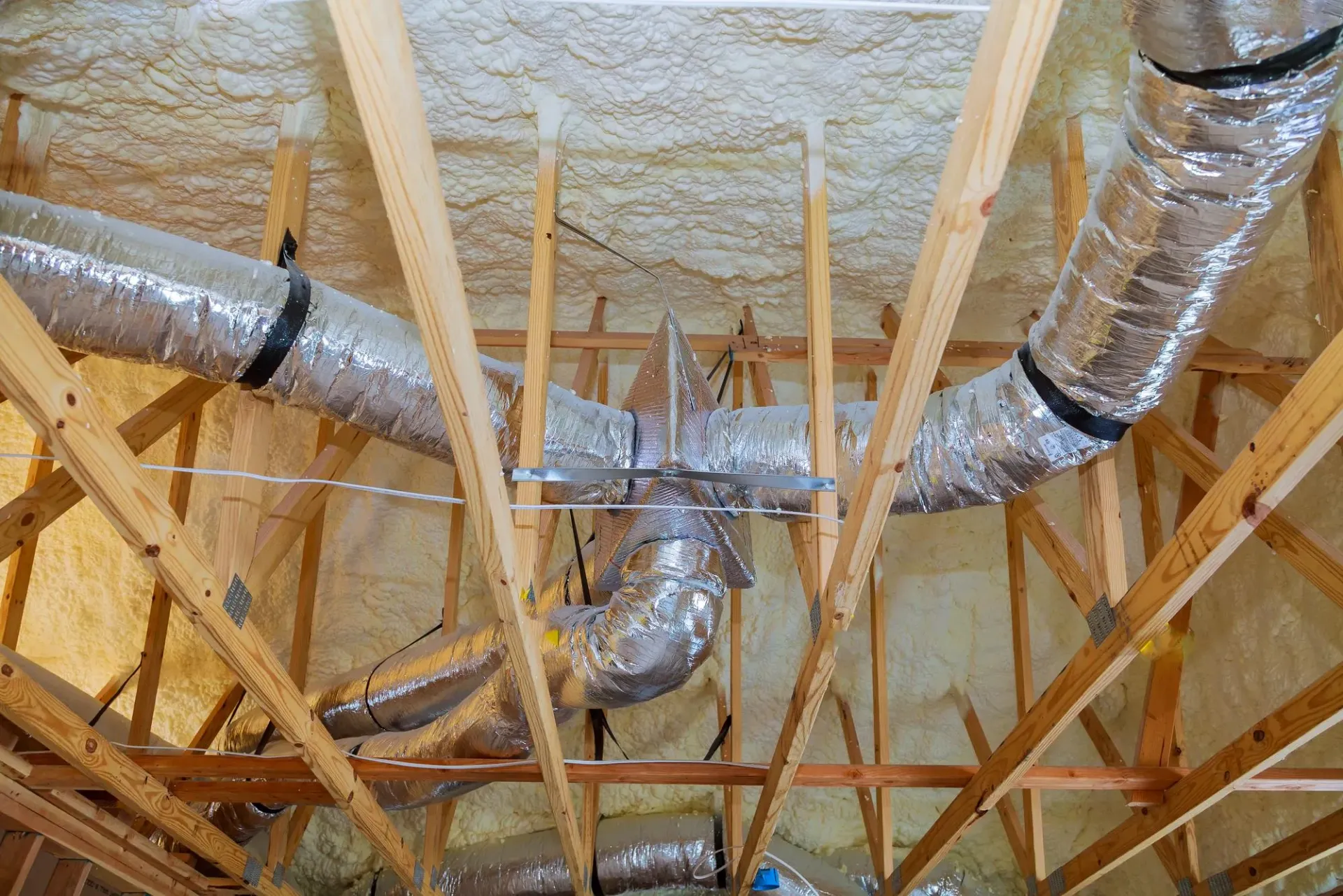 An interior view of a wooden attic frame with foam insulation between beams and a square skylight open to the sky.