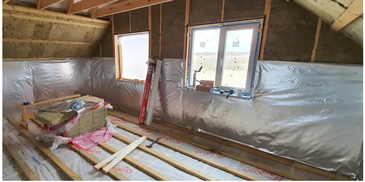 A person in a hard hat standing on a ladder and spraying white foam insulation onto an unfinished interior wall.