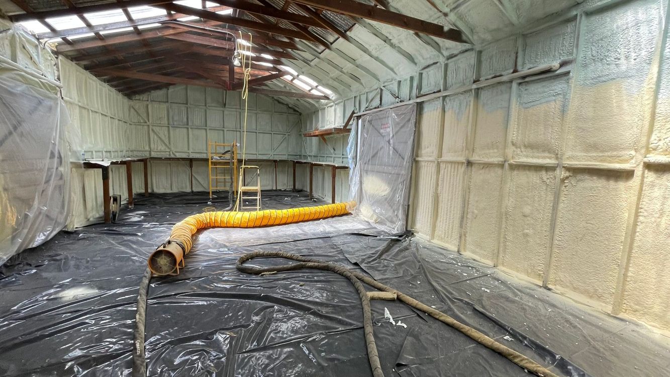 Inside a building, walls and ceilings are coated with tan spray foam insulation. A yellow ventilation hose lies on floor.