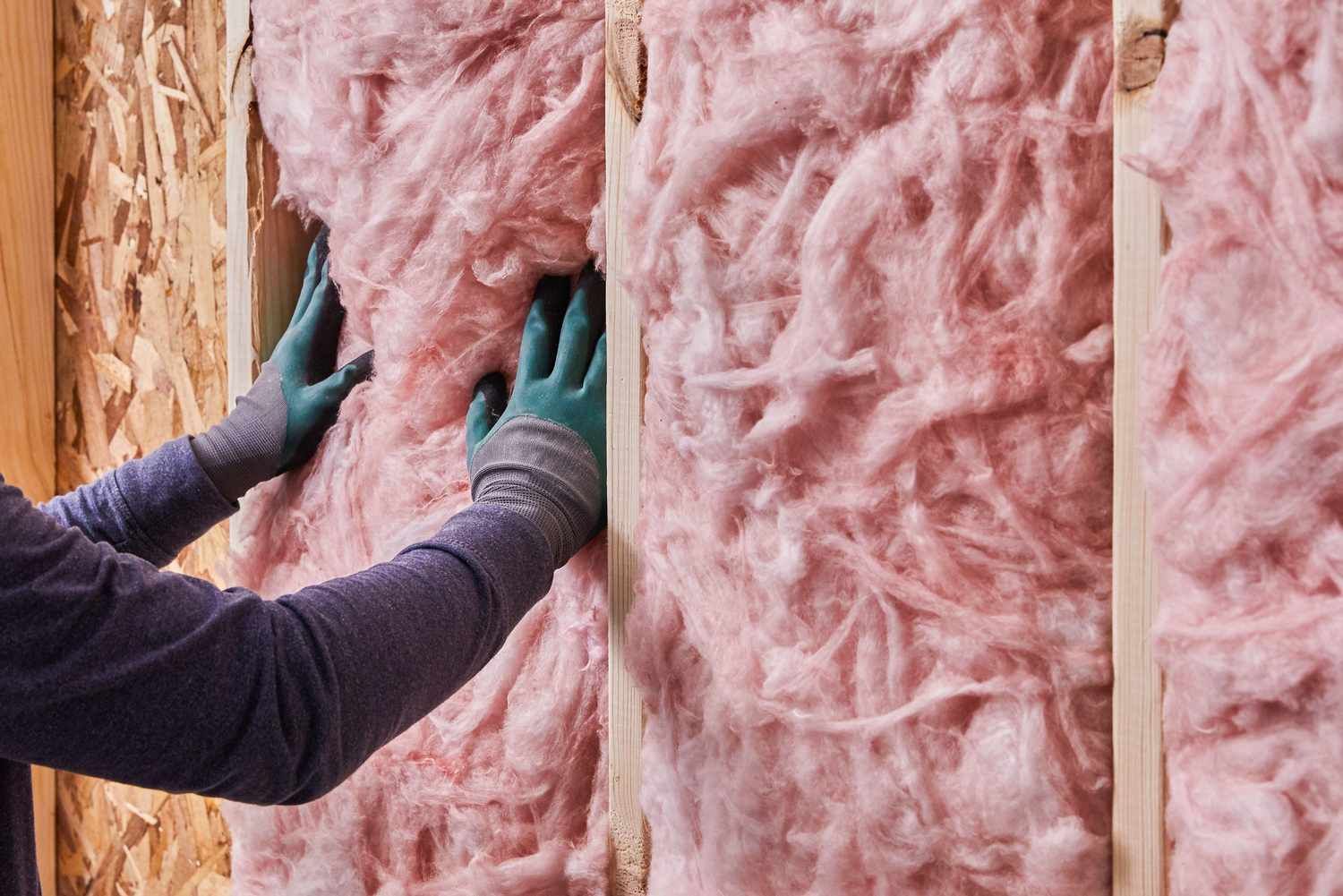 A person in a hard hat standing on a ladder and spraying white foam insulation onto an unfinished interior wall.