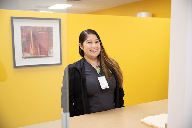 A smiling person in scrubs and a black jacket behind a reception desk in a room with yellow walls and framed art.