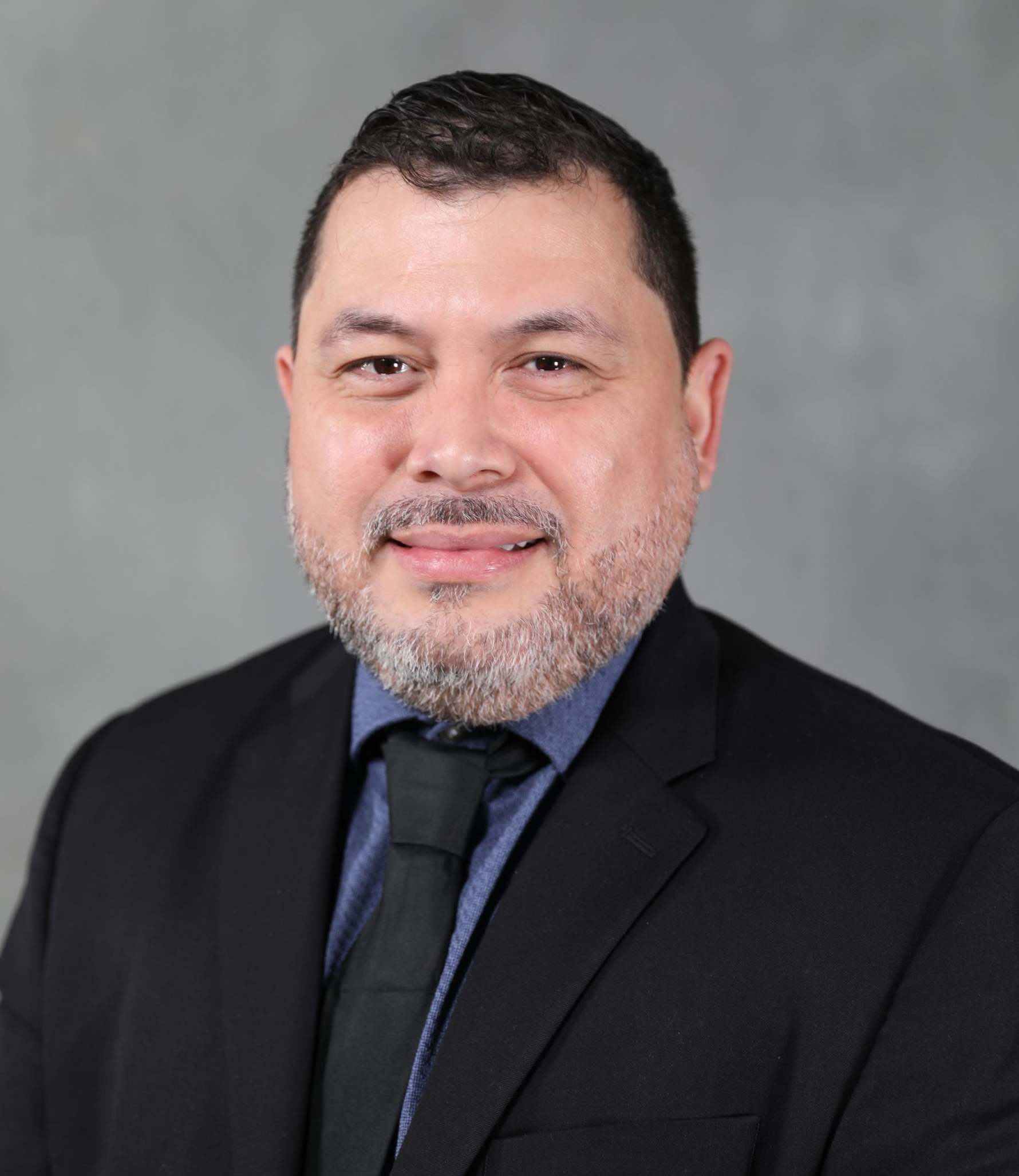 A person with a short beard wearing a dark suit, blue shirt, and black tie against a gray background.