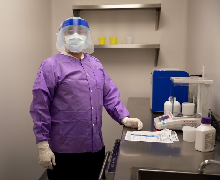 A person in a purple lab coat, face mask, and face shield stands in a laboratory next to equipment and a stainless counter.