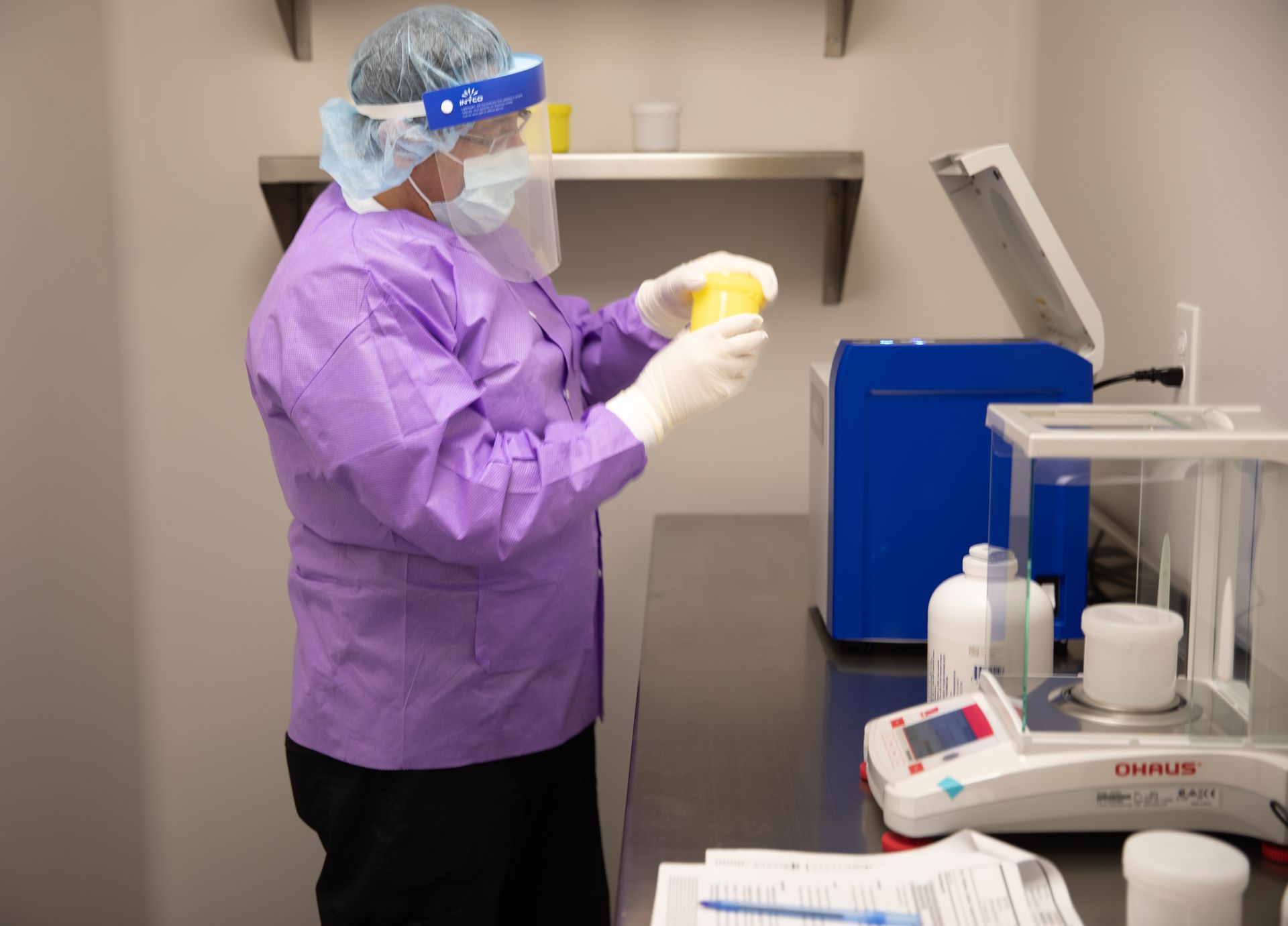 A person in a purple lab coat, face shield, and gloves holds a yellow container in a laboratory next to a scale.