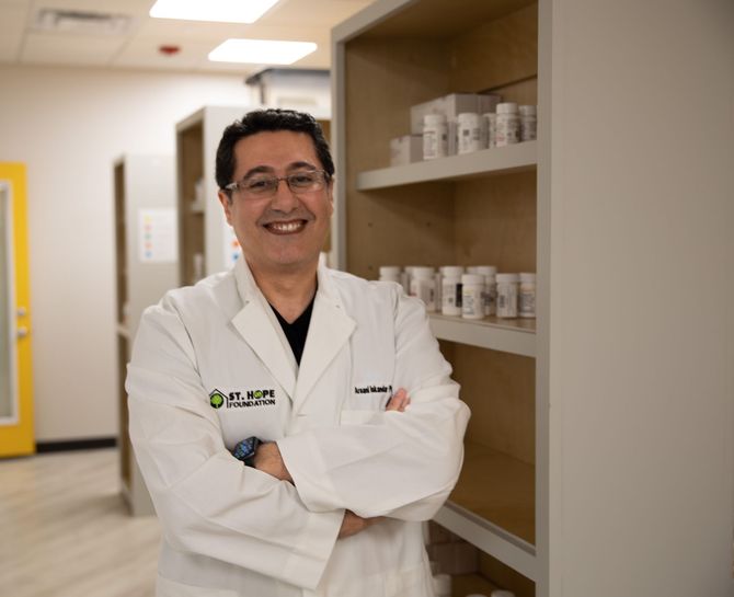A smiling person in a white lab coat stands with arms crossed in a room with shelves of medicine bottles.