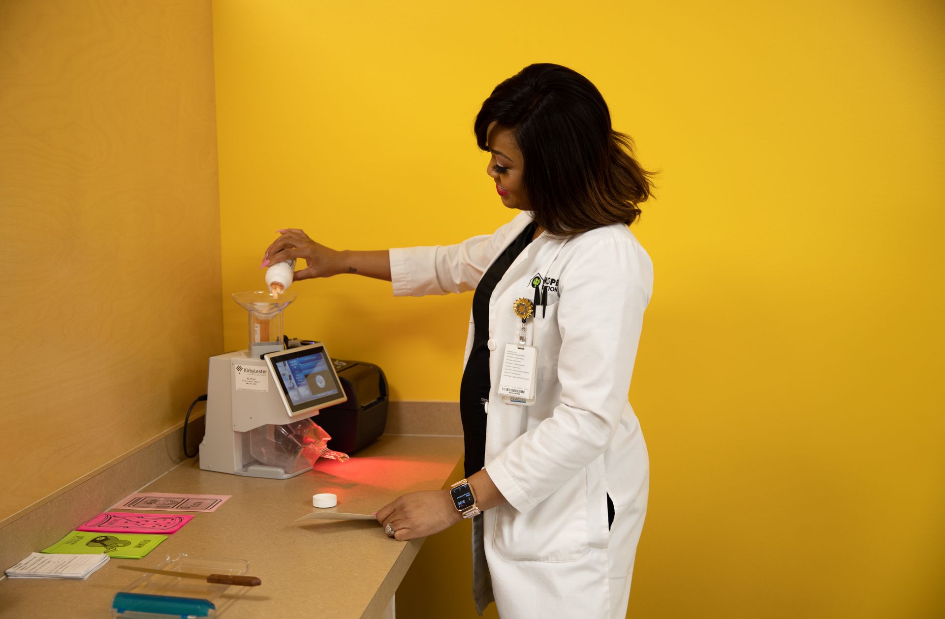 A healthcare worker in a white coat uses a pill counting machine in a room with bright yellow walls.