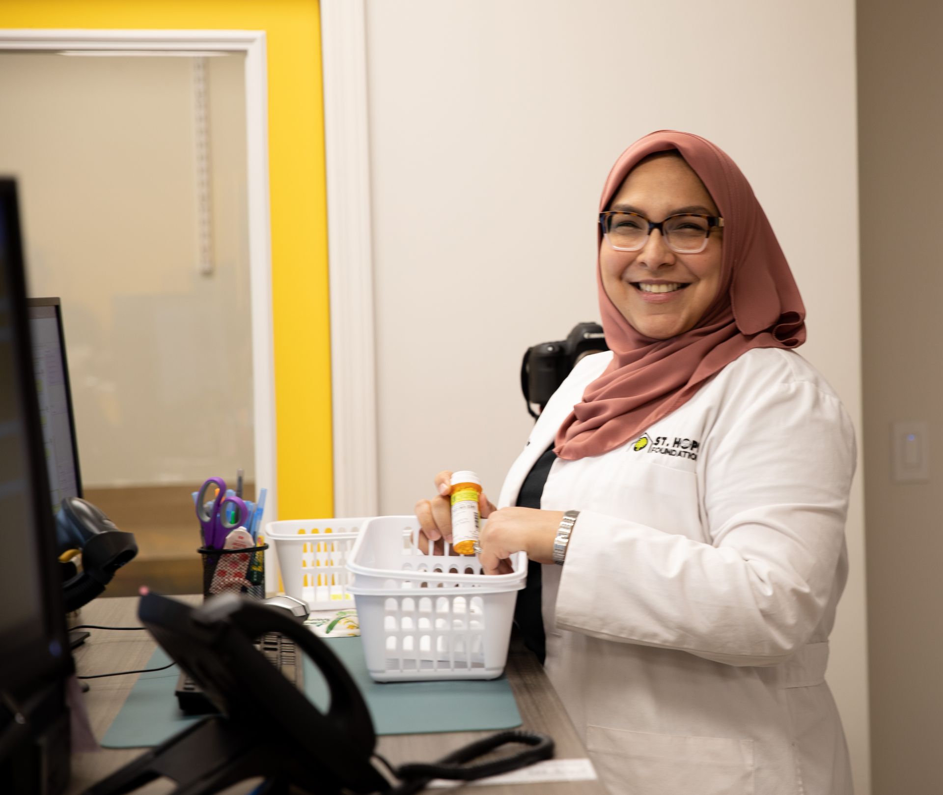 A pharmacist wearing a lab coat and hijab smiles while sorting a medication bottle into a white basket on a desk.