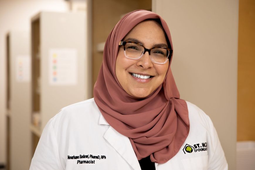 A smiling pharmacist in a white coat and dusty rose hijab, standing in a clinical setting.