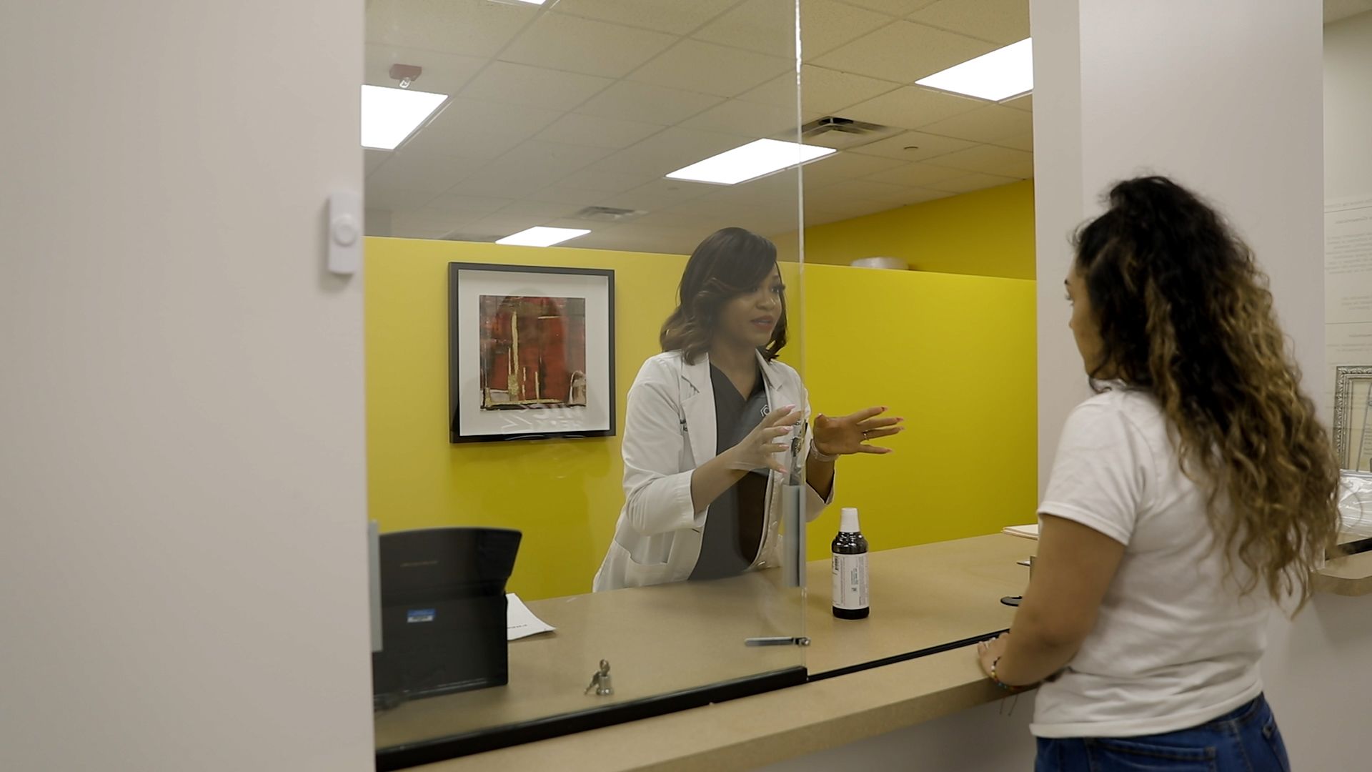 A pharmacy staff member in a white coat speaks to a customer across a counter with a protective glass partition.
