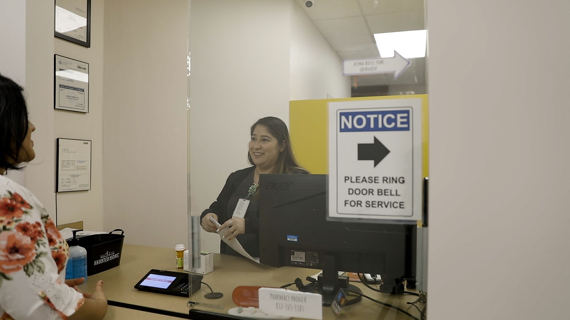 A person at a reception desk speaks to an employee behind a glass partition, which features a notice to ring the doorbell.