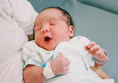 Newborn baby yawning, held in someone's arms, wearing a hospital wristband.
