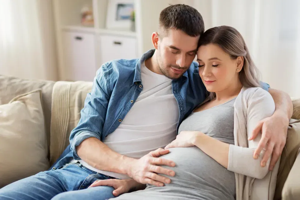 Pregnant woman and man embracing on a sofa, touching her belly in a living room.
