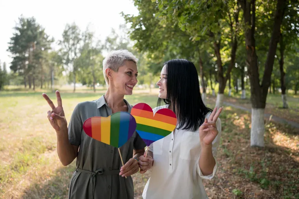 Two people holding rainbow heart signs, smiling, giving peace signs, in a park.