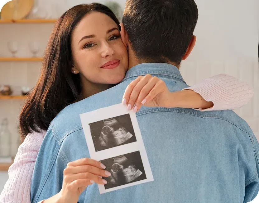Woman hugs man, holding sonogram images. They are in a well-lit kitchen.