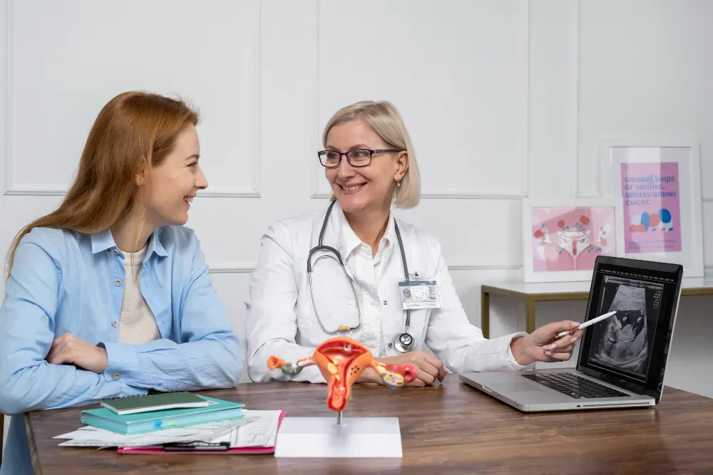 Pregnant person in blue gown consults with a doctor in a medical office; doctor holds clipboard.
