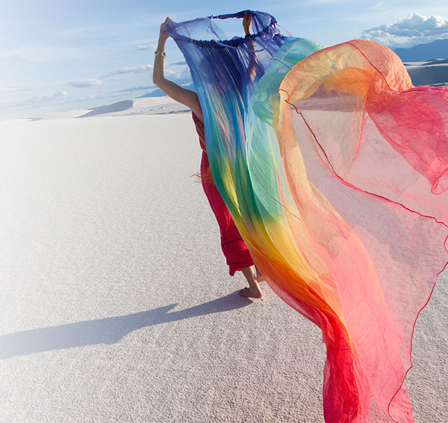 Woman in red holds a rainbow scarf high in a white desert, arms raised.