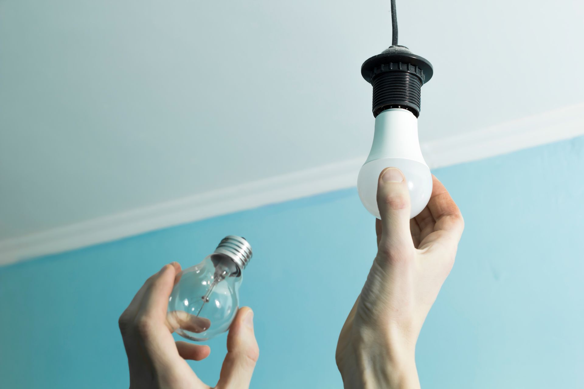 A person holding an LED light bulb up on a ceiling with one hand and a traditional bulb in the other