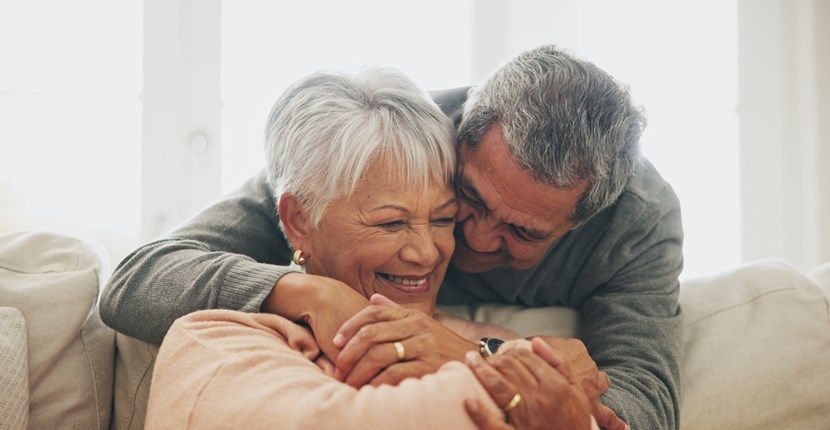 An elderly couple is hugging each other on a couch.