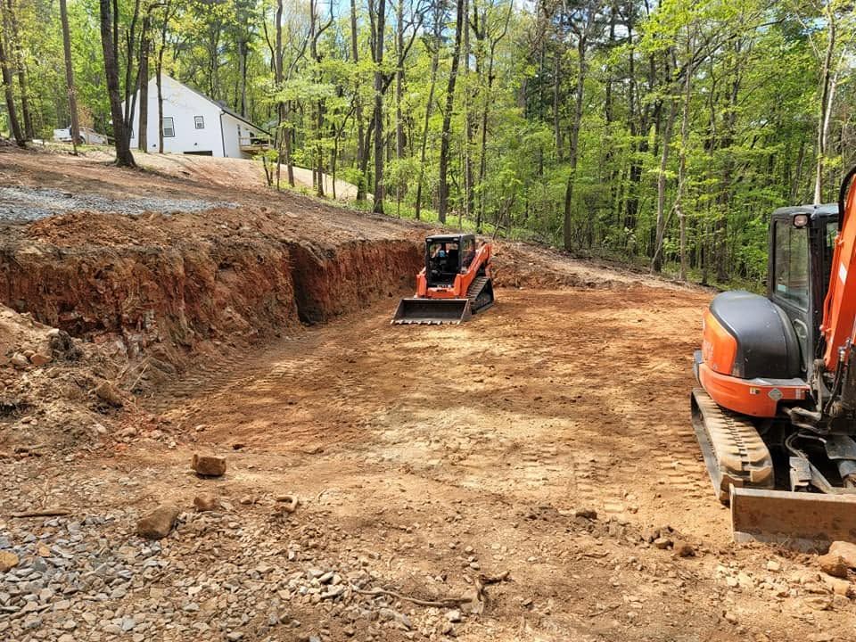 Orange excavators grading a dirt road on a wooded hillside near a white house