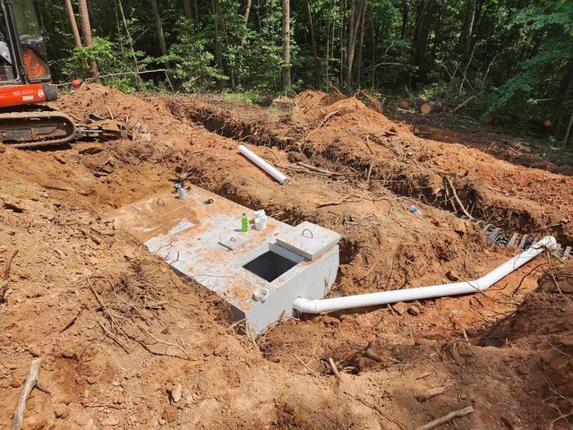Excavated dirt site with a concrete septic tank and white PVC pipes in a wooded area.