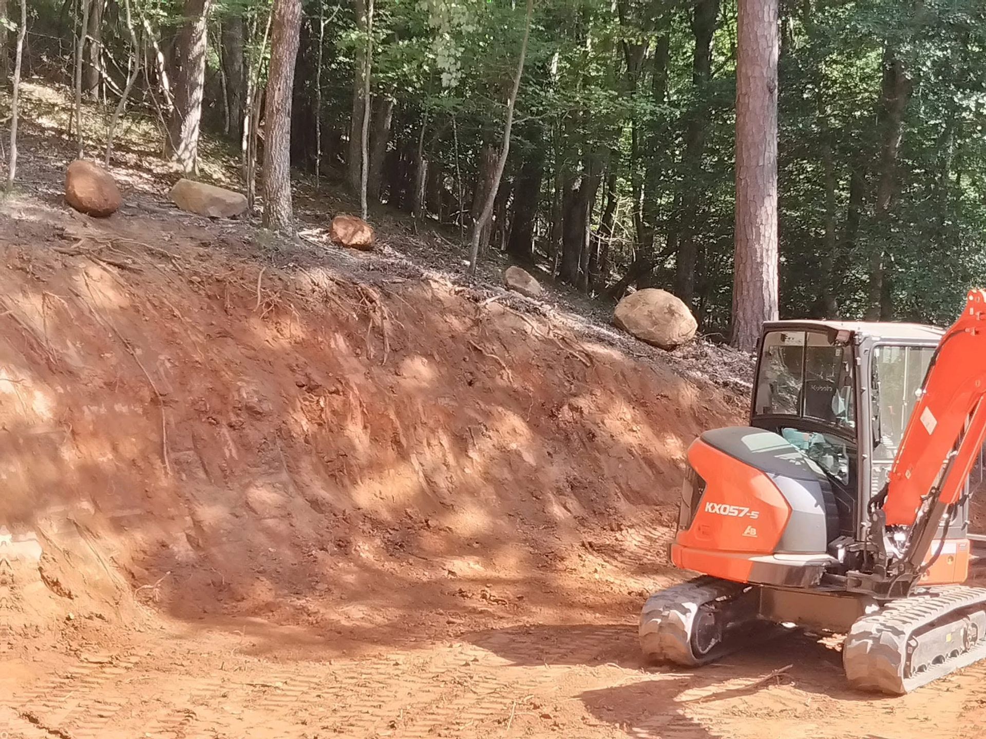 Excavator working on a dirt slope in a wooded area with large rocks nearby