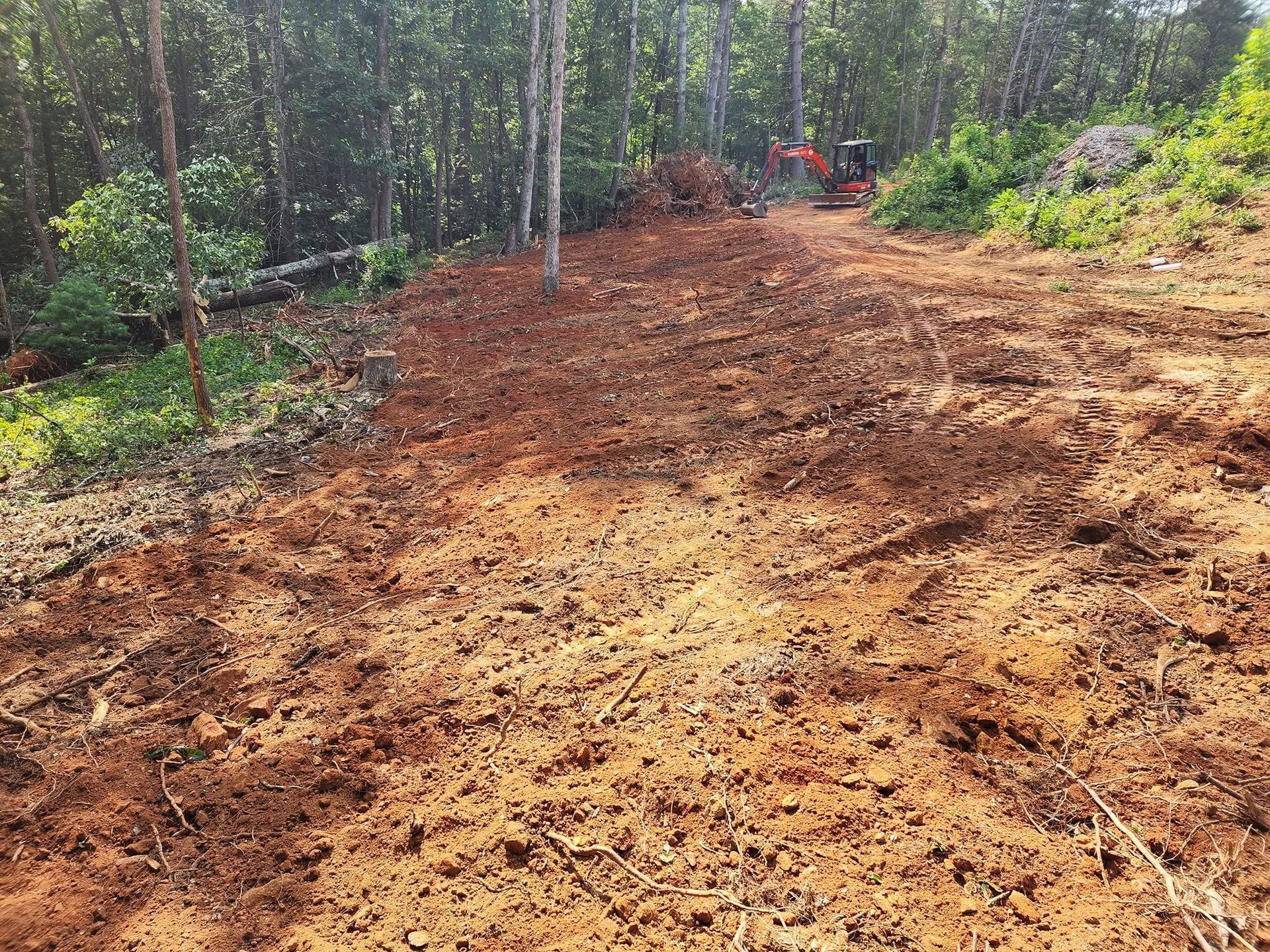 Dirt road under construction through a wooded area, with a small excavator in the distance.