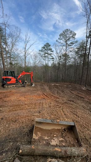 Orange excavator clearing a dirt lot in a wooded area, with a bucket in the foreground.