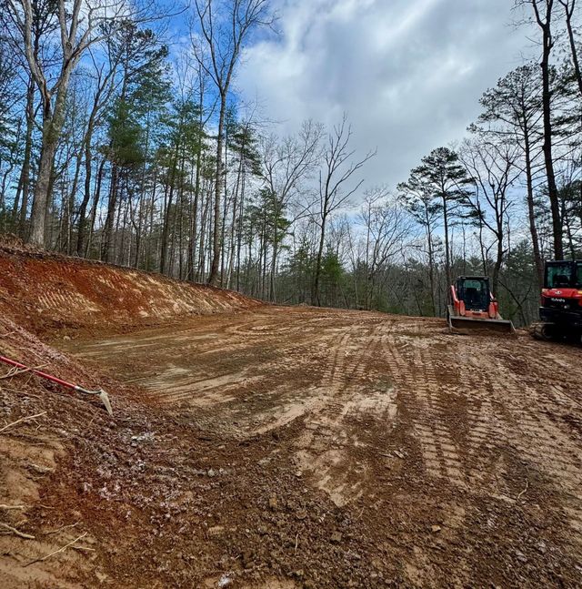 Cleared dirt lot in a wooded area with two construction machines on the right side.