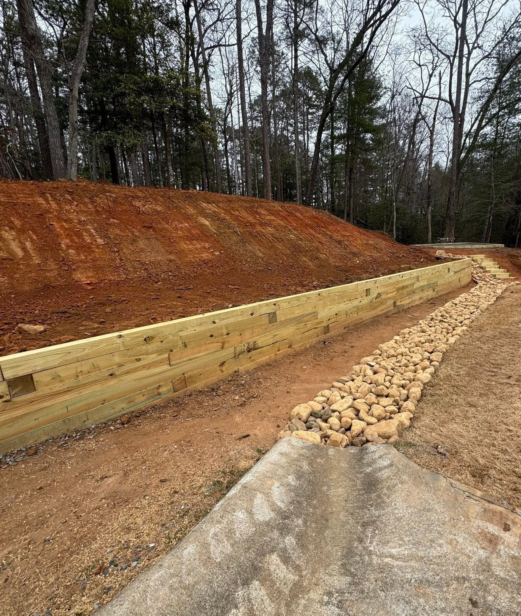 Gravel path beside a wooden retaining wall and reddish dirt embankment in a wooded area