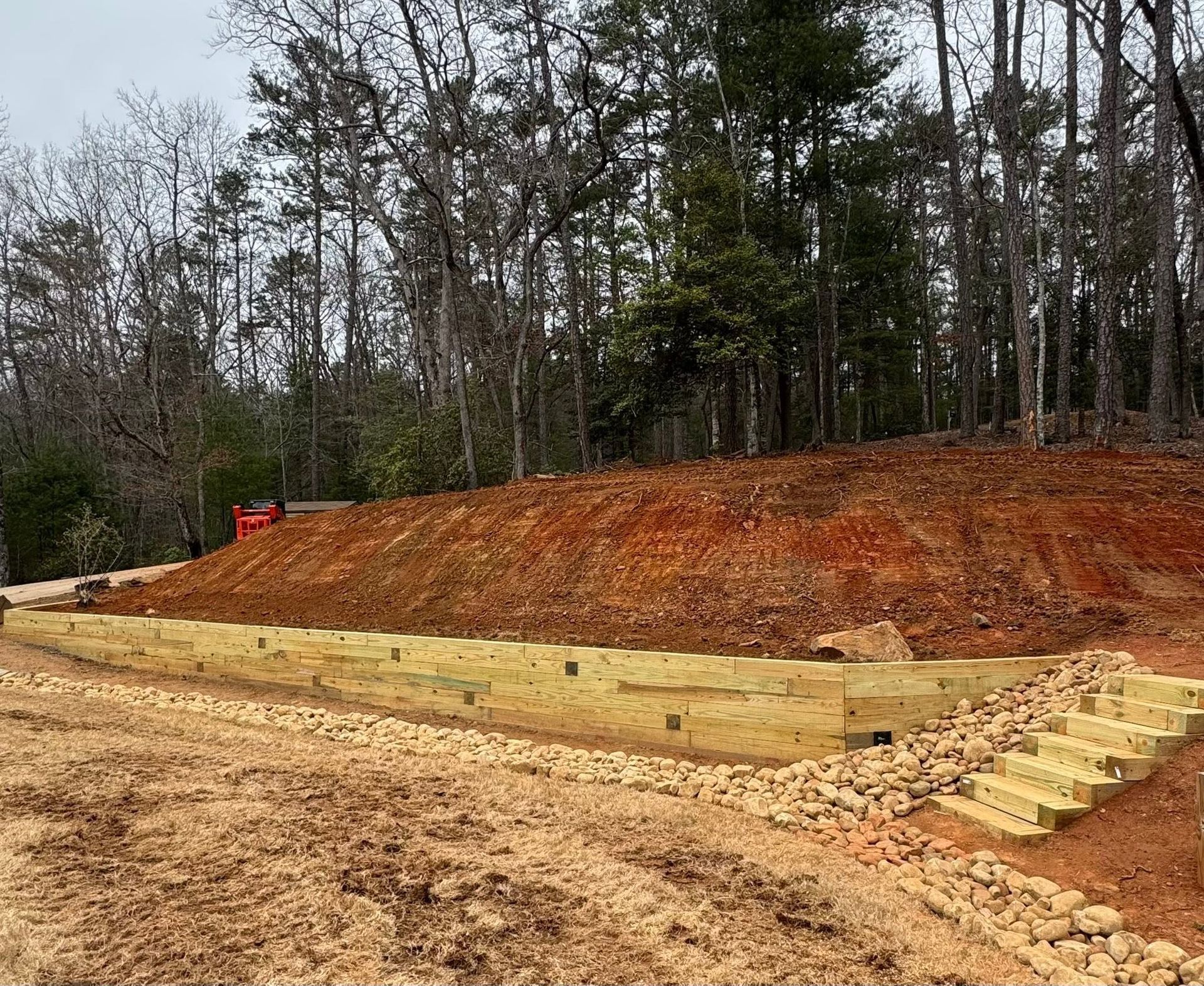 Construction site with a wooded slope, yellow retaining wall, and stepped stone stairs.