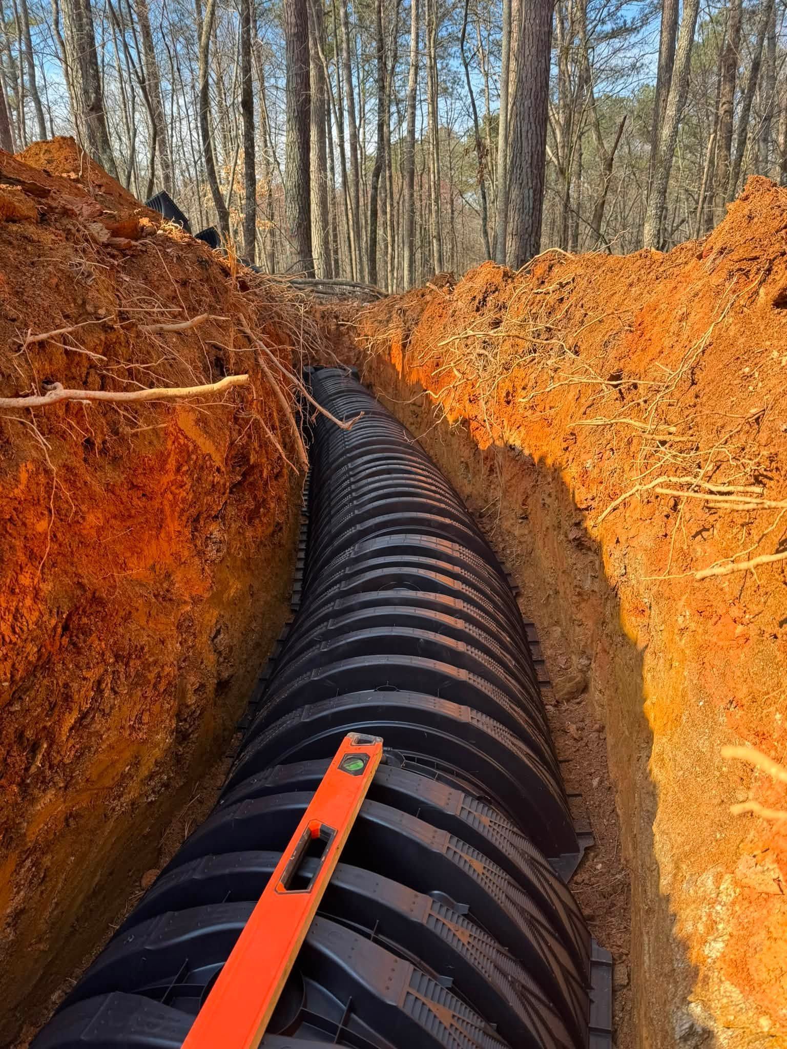 Black drainage pipe in a narrow trench with orange dirt, lined by trees in the background