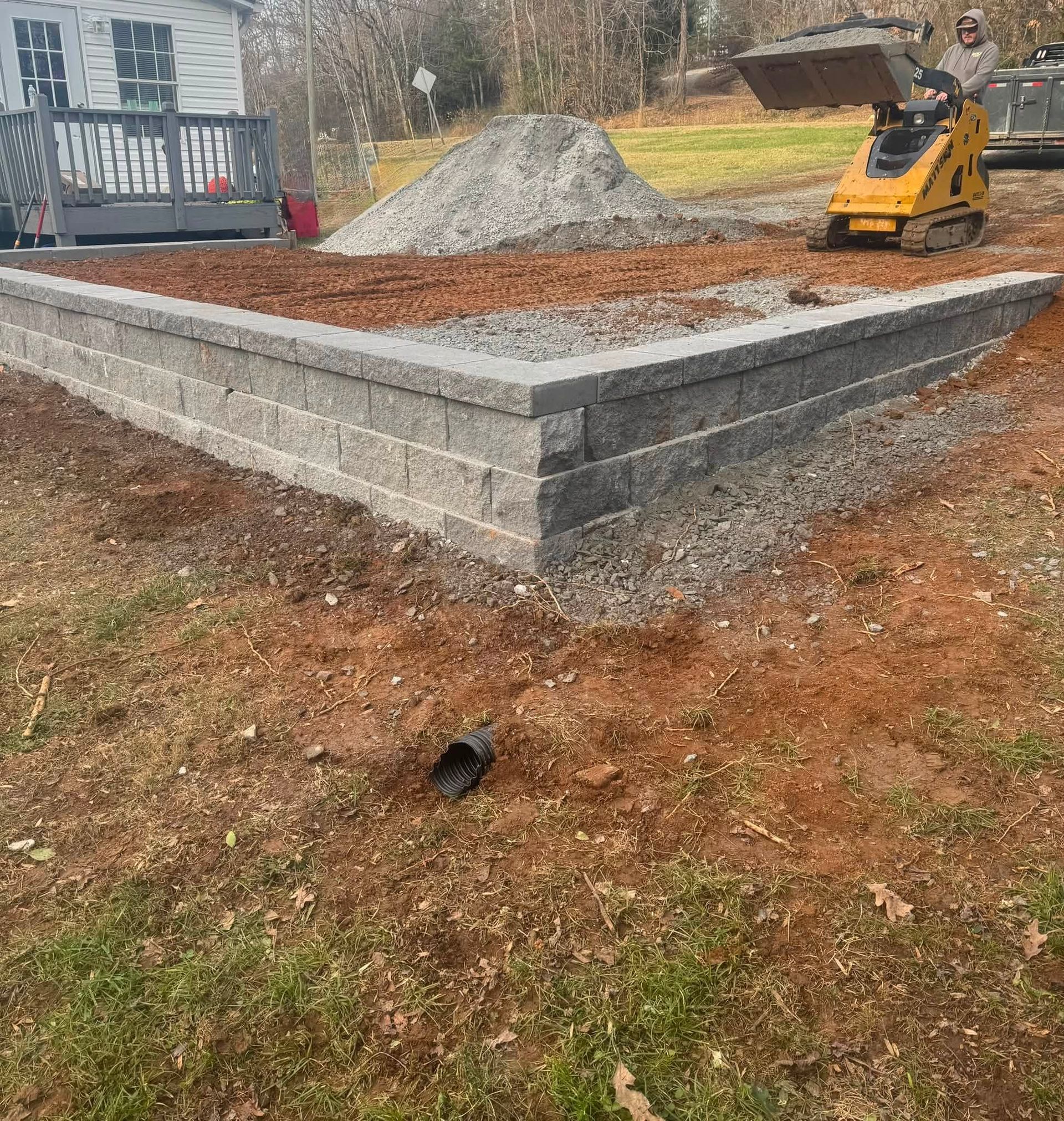 Retaining wall under construction with gravel, excavator, and a black pipe in a yard