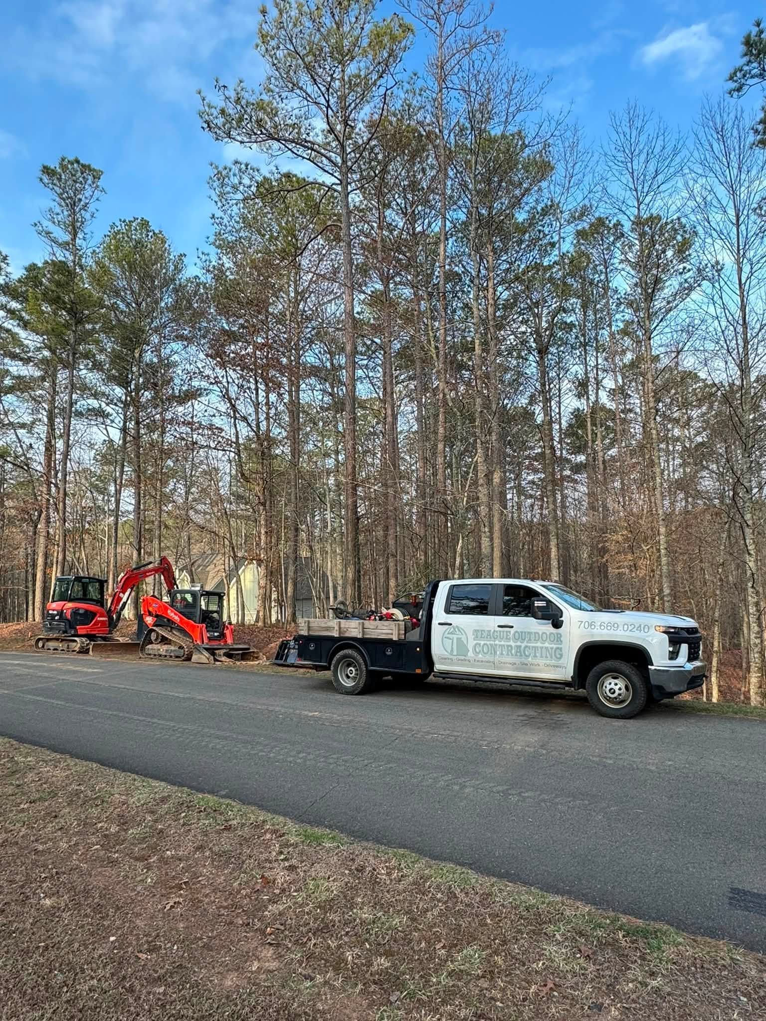 White pickup truck with trailer and red equipment parked on a road beside a wooded area under a blue sky