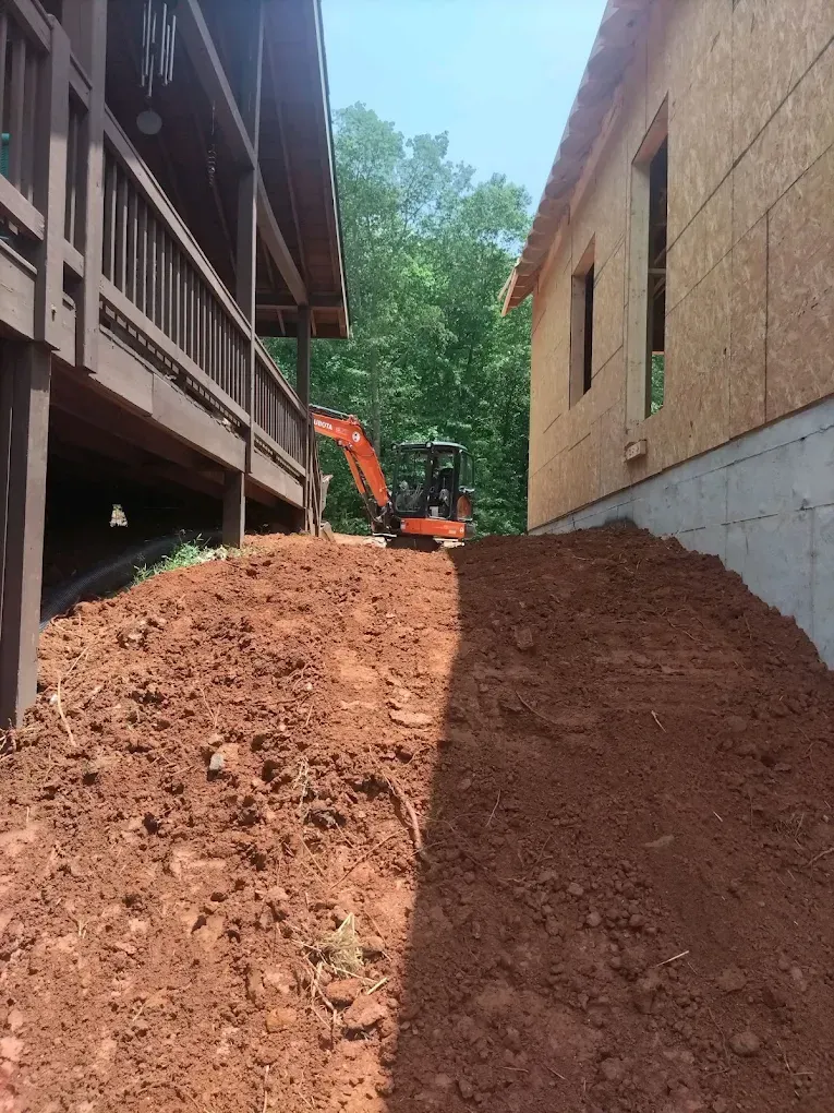 Construction equipment on a dirt slope between two buildings, with an excavator in the distance.