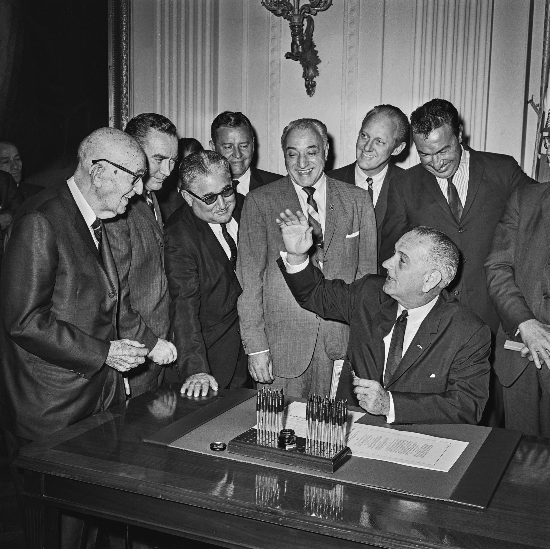 Group of men gathered around a seated man at a desk in a formal room, one raising a glass in a toast.