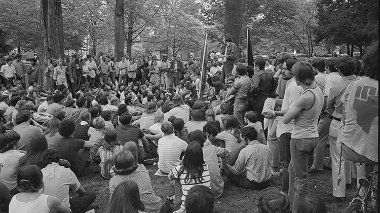 Crowd seated outdoors in a park, listening to a speaker near a small stage or gathering area.