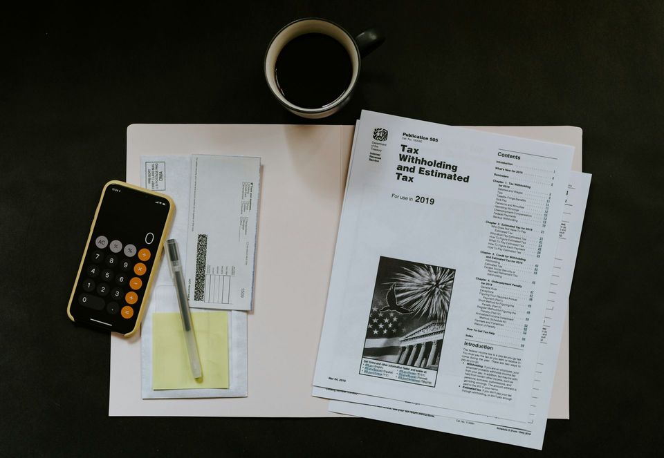 Desk with scattered papers, calculator, pen, tape roll, and a yellow sticky note on a dark surface.