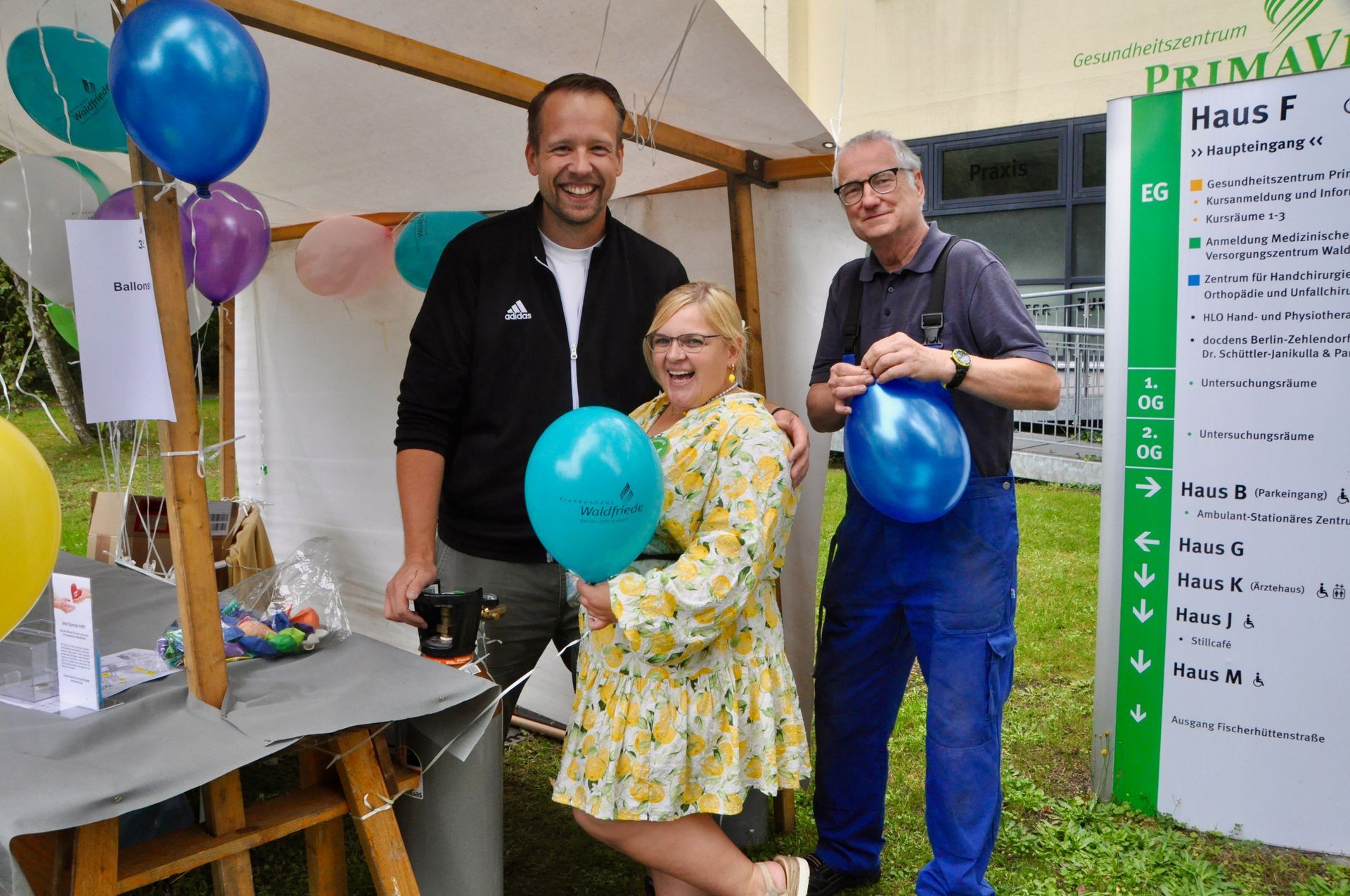 Drei lächelnde Personen stehen neben einem Zelt mit Luftballons; ein Schild mit der Aufschrift PrimaVera Haus F ist zu sehen.
