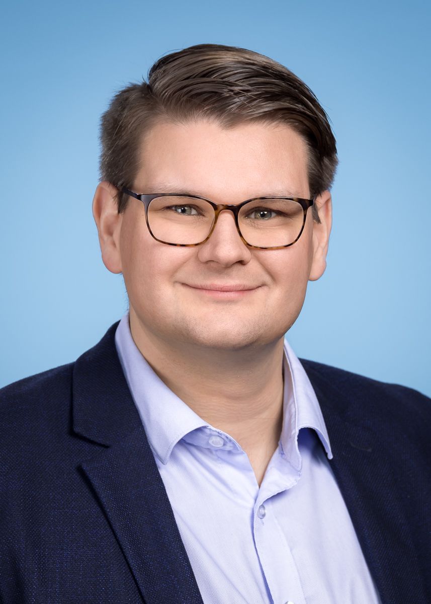 Ein Mann mit Brille und Blazer lächelt vor einem blauen Hintergrund.