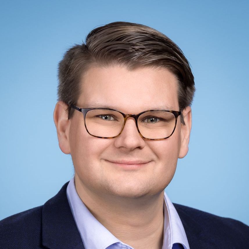 Mann mit Brille und dunkelblauem Blazer lächelt vor blauem Hintergrund.