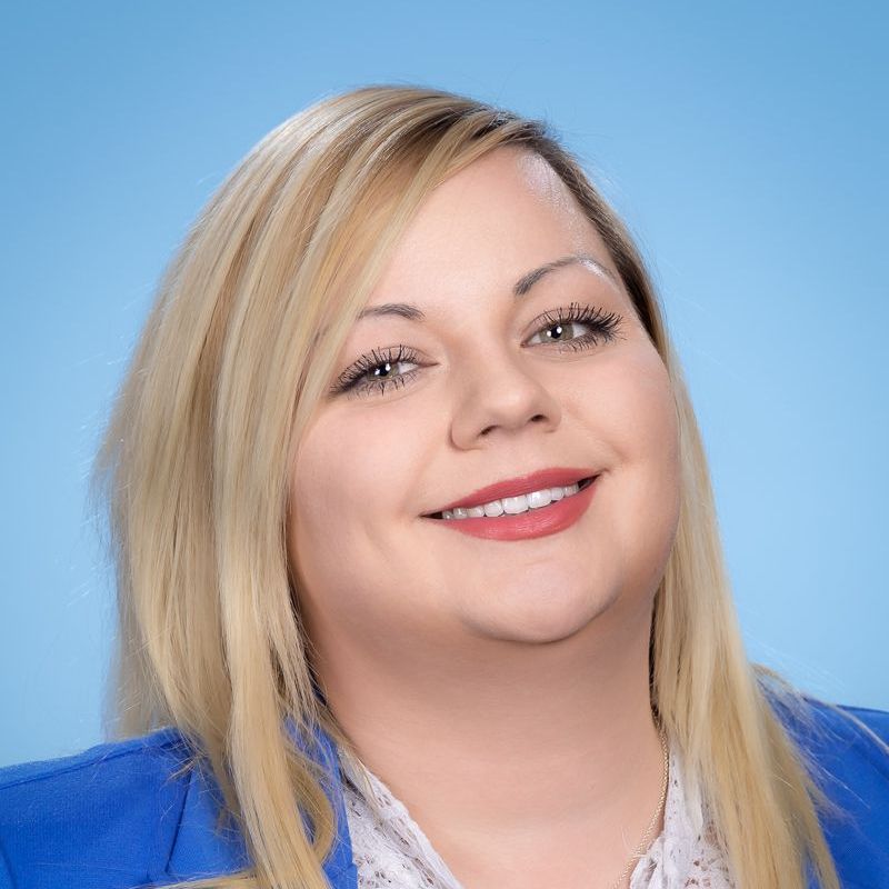 Eine Frau mit blondem Haar trägt eine blaue Jacke und lächelt.
