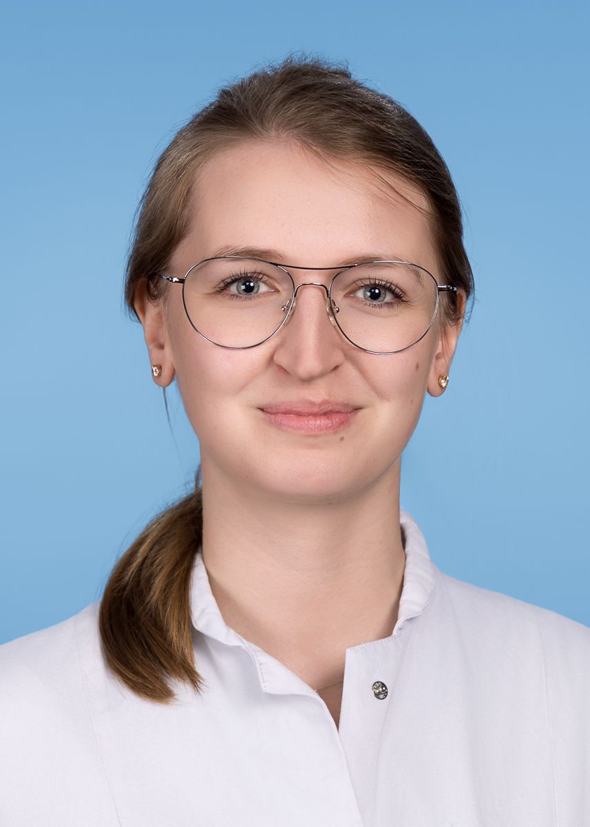 Frau mit Brille, lächelnd, hellbraunes Haar zu einem tiefen Pferdeschwanz gebunden, trägt einen weißen Mantel, vor hellblauem Hintergrund.
