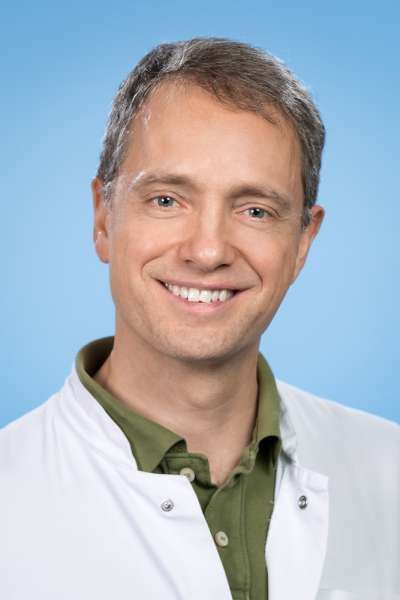 Ein Mann in einem weißen Laborkittel und einem grünen Hemd lächelt in die Kamera.